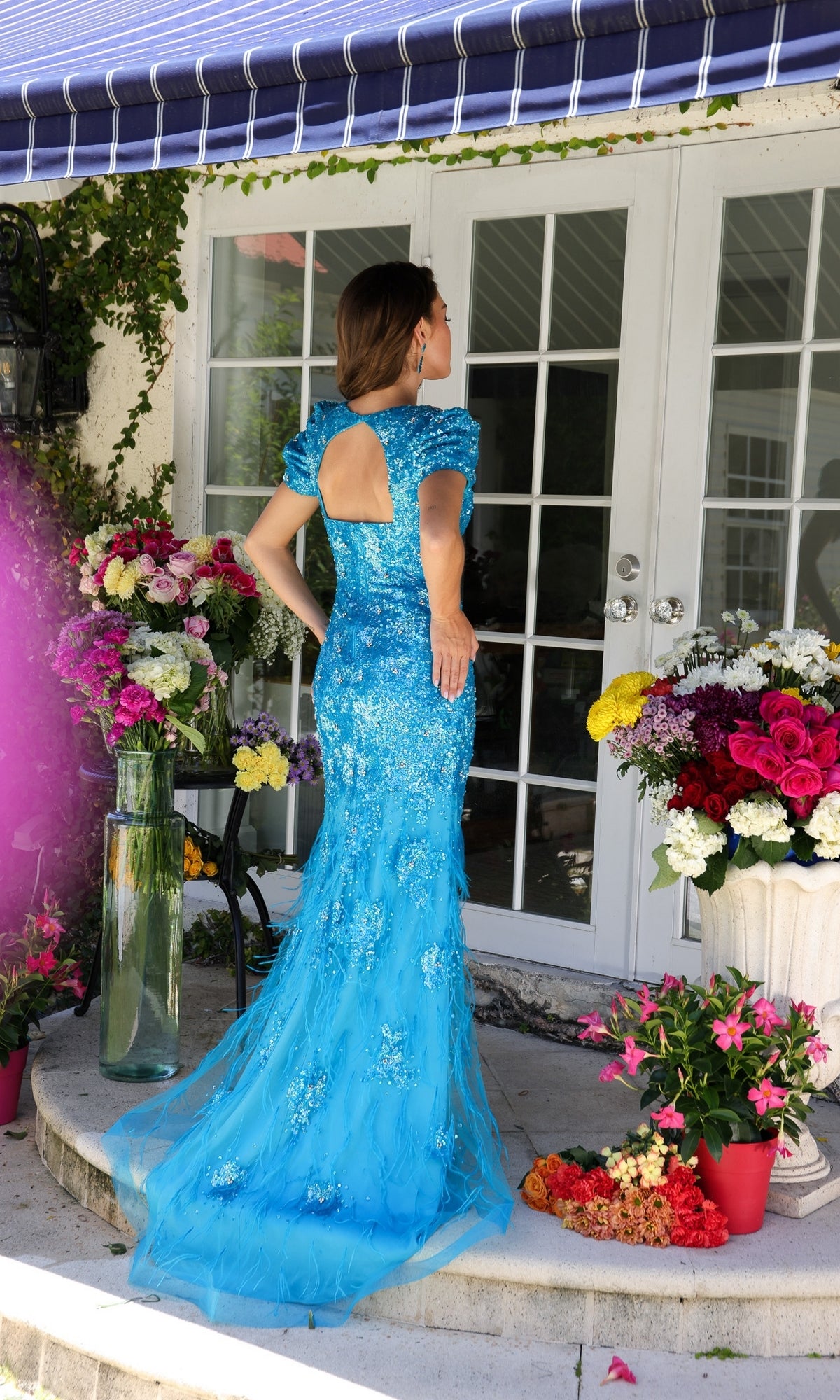 A woman in the Ava Presley 39552 Feather Short-Sleeve Prom Gown, blue, stands on a porch amid colorful flowers, facing away toward glass doors.